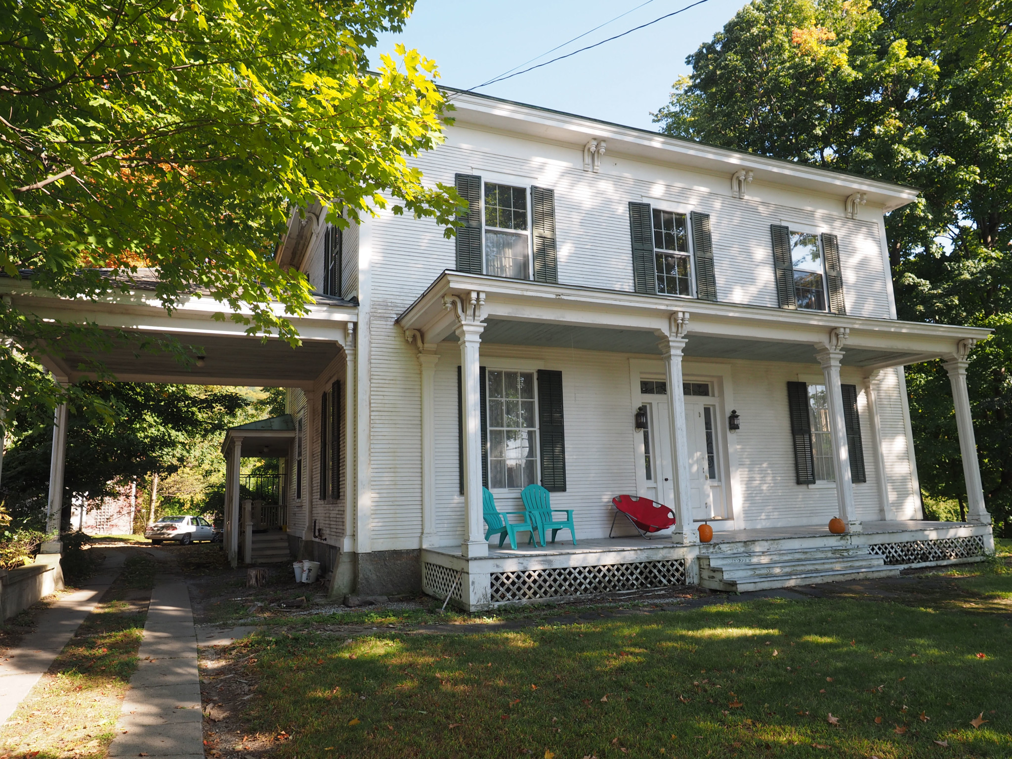 White historic house with front porch and colorful chairs, surrounded by trees.