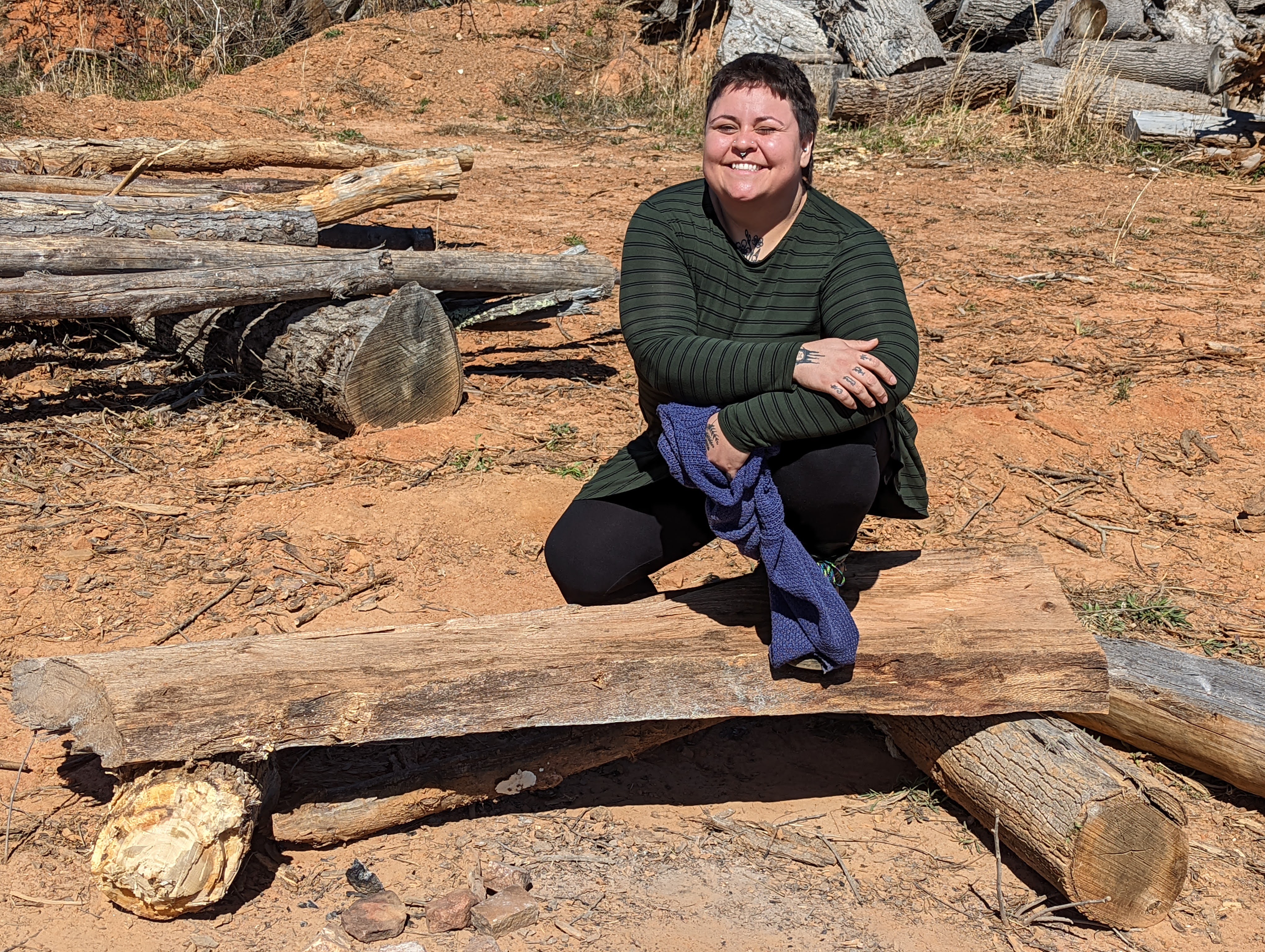 Person sitting on a log outdoors with a smile, surrounded by logs and natural scenery.