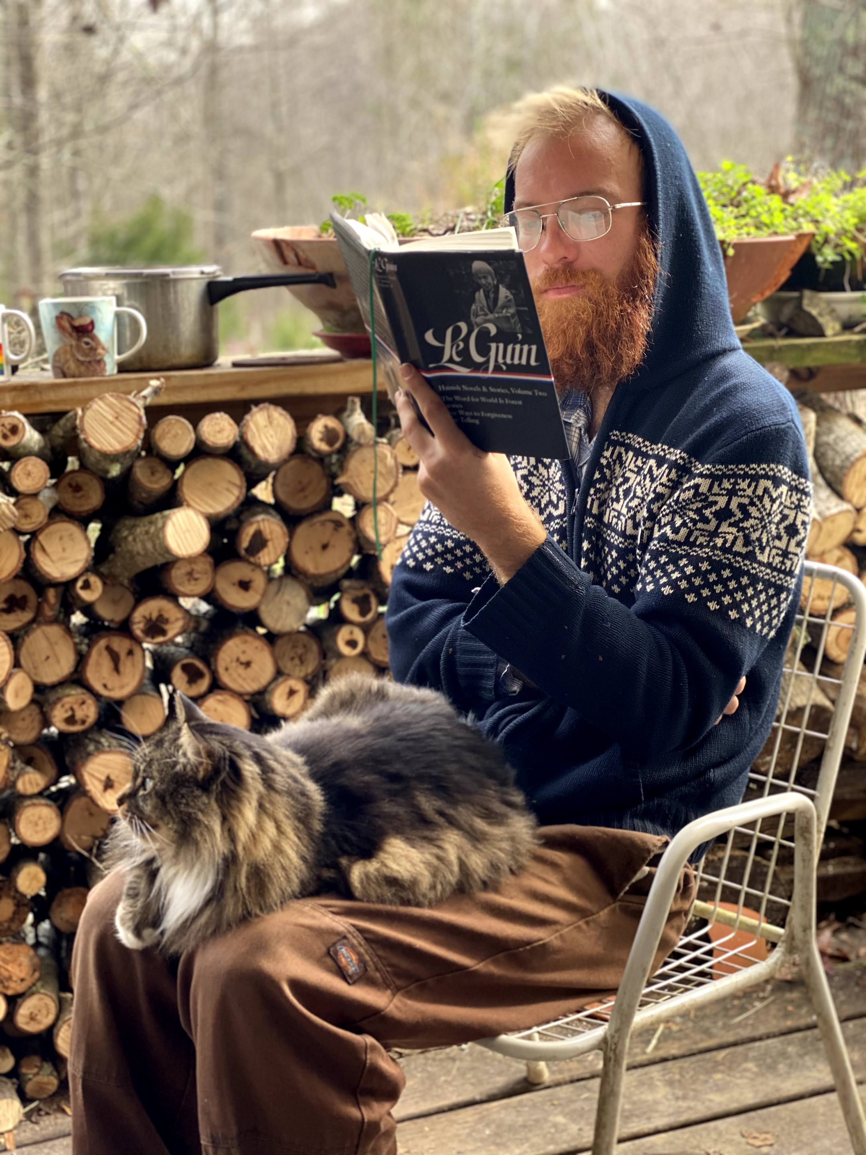 Man with glasses reading a book outdoors with a cat on his lap.