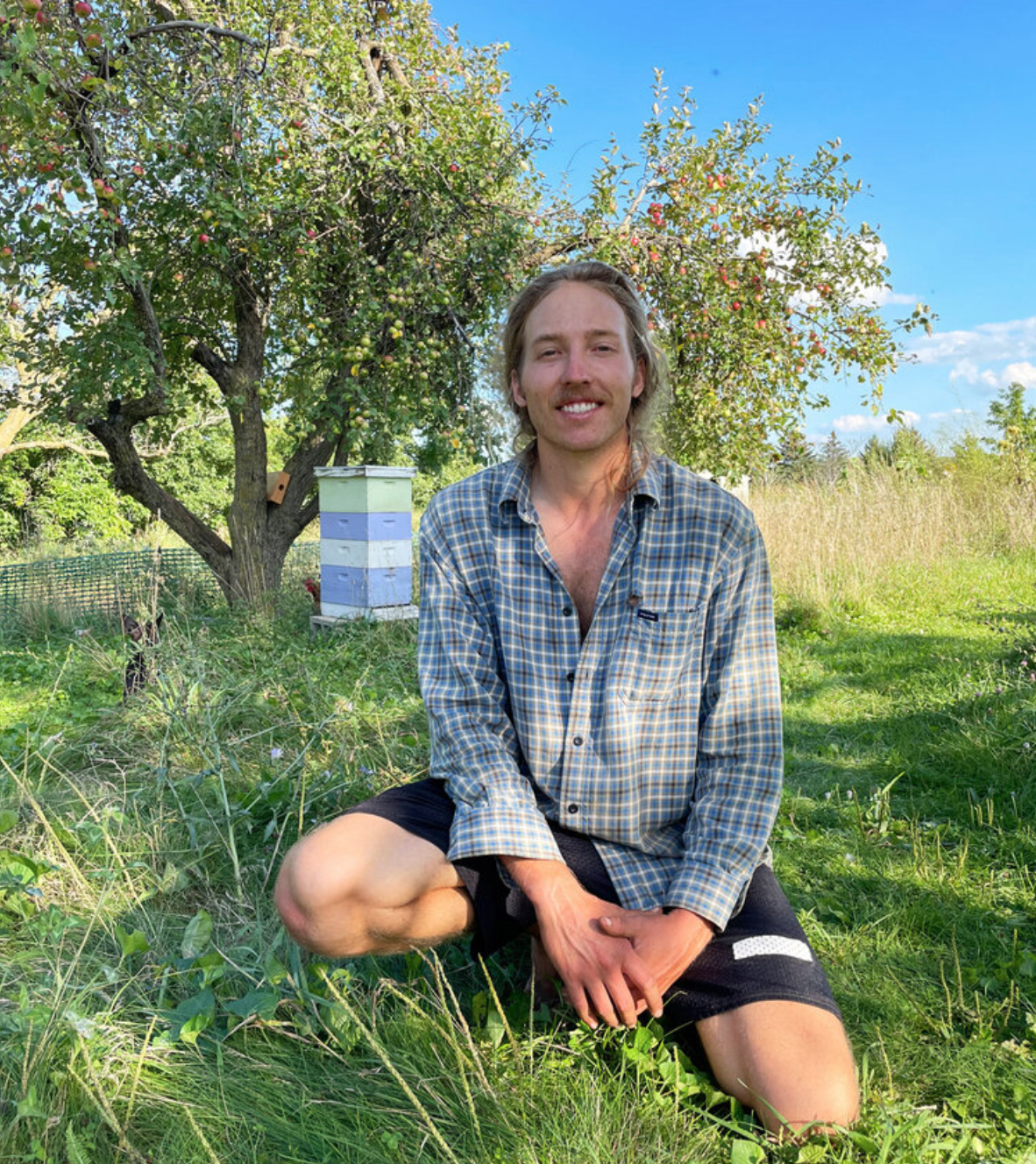 Person smiling in orchard with beehives in background at Greenbush Growing Cooperative.