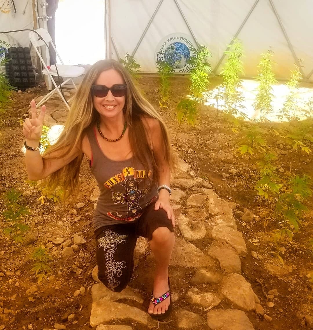 Woman smiling in a greenhouse with cannabis plants, making a peace sign.