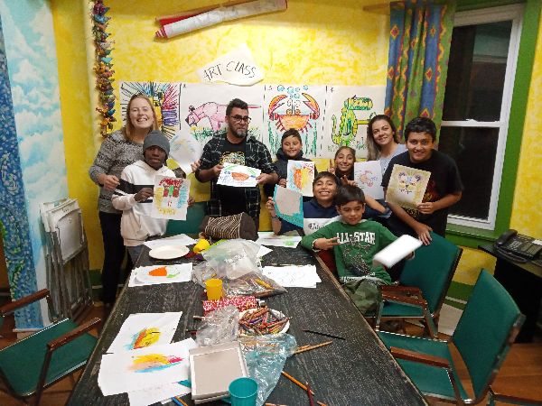 Group of children and adults holding art projects in a colorful classroom.