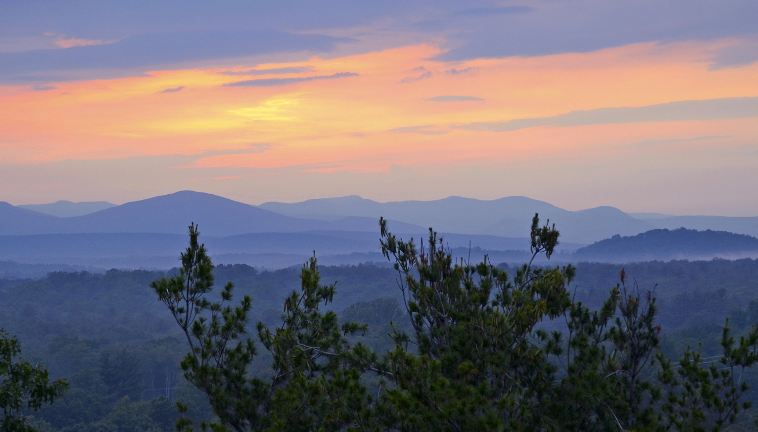 Beautiful mountain landscape during sunset with layered mountain ranges and trees in foreground.
