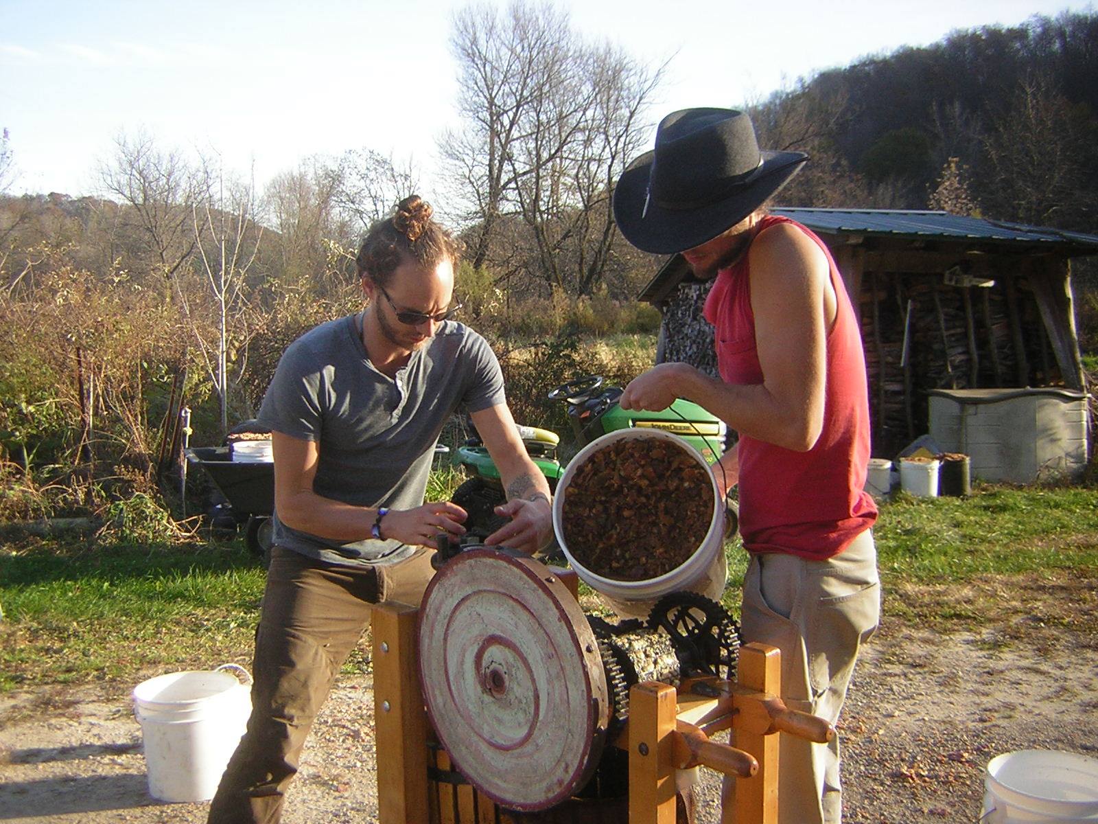 Two women operating a manual seed grinder outdoors in a permaculture setting.