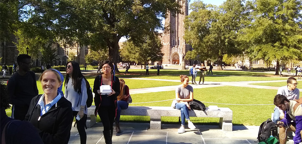 People gathering in a park near Durham Bruderhof House with trees and historic buildings.