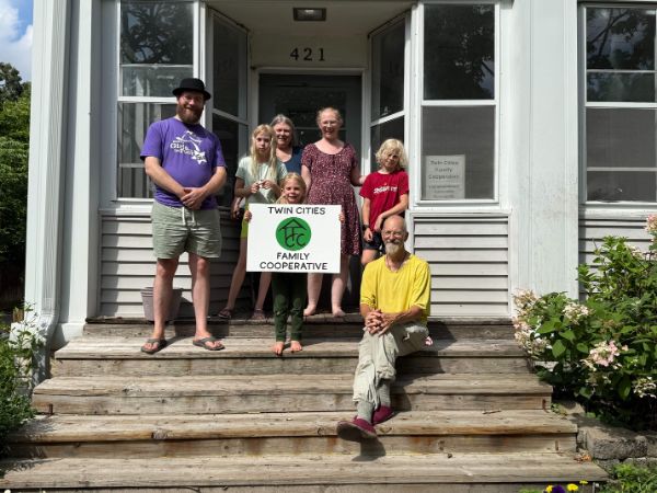 Group of diverse people holding a Twin Cities Family Cooperative sign in front of a house.