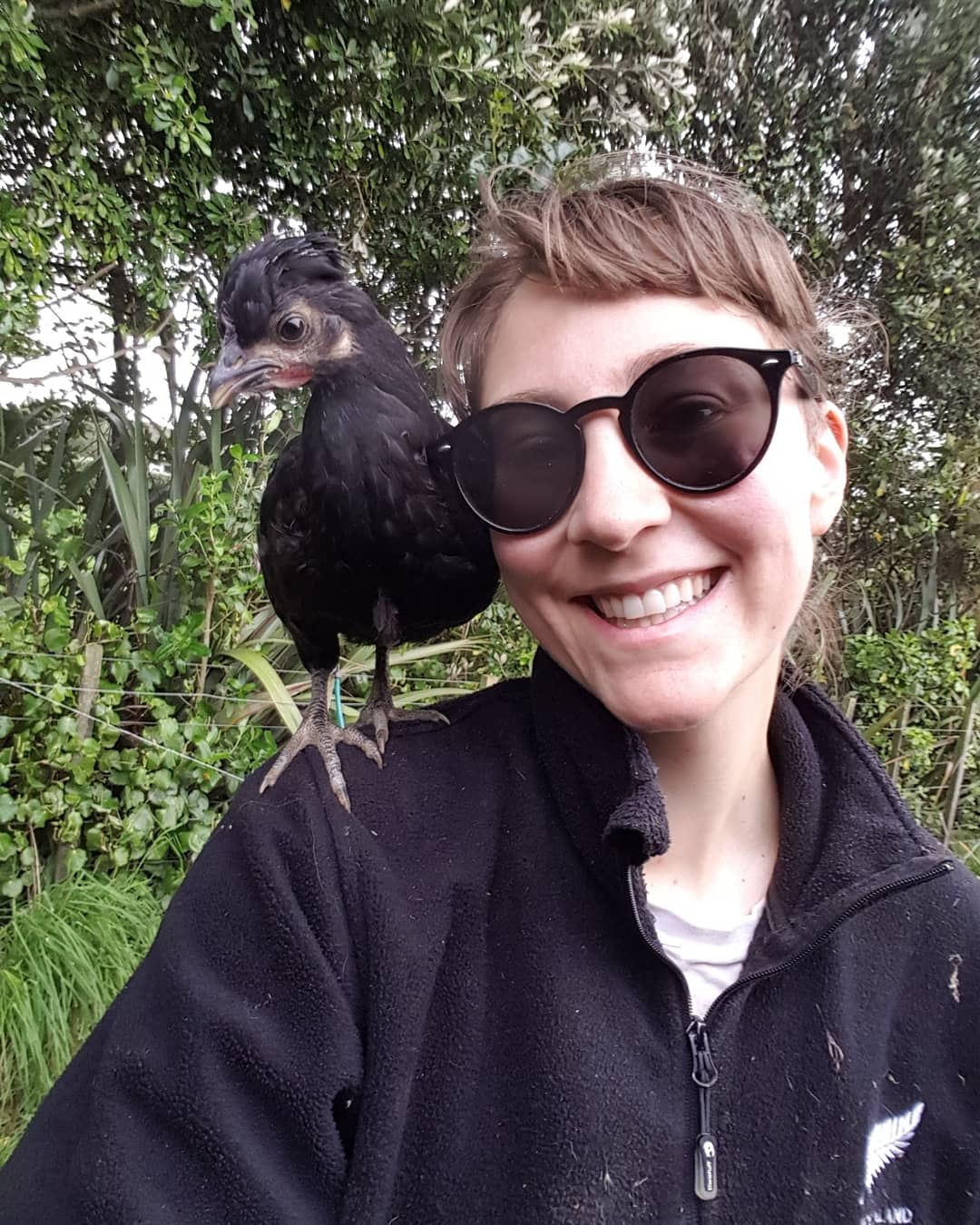 Young person with sunglasses smiling, bird perched on shoulder in nature setting.
