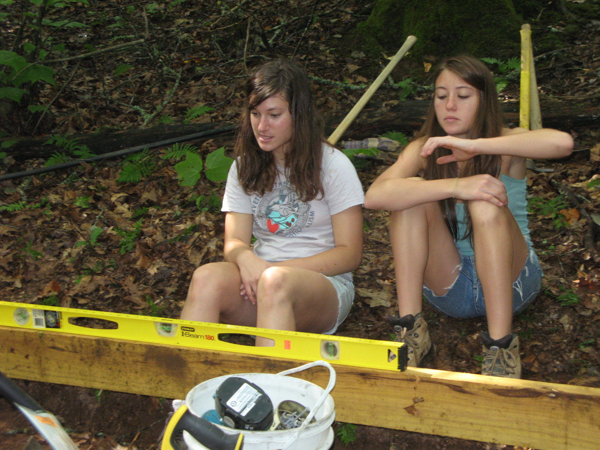 Two young girls sitting on the ground in a wooded area during outdoor activity.