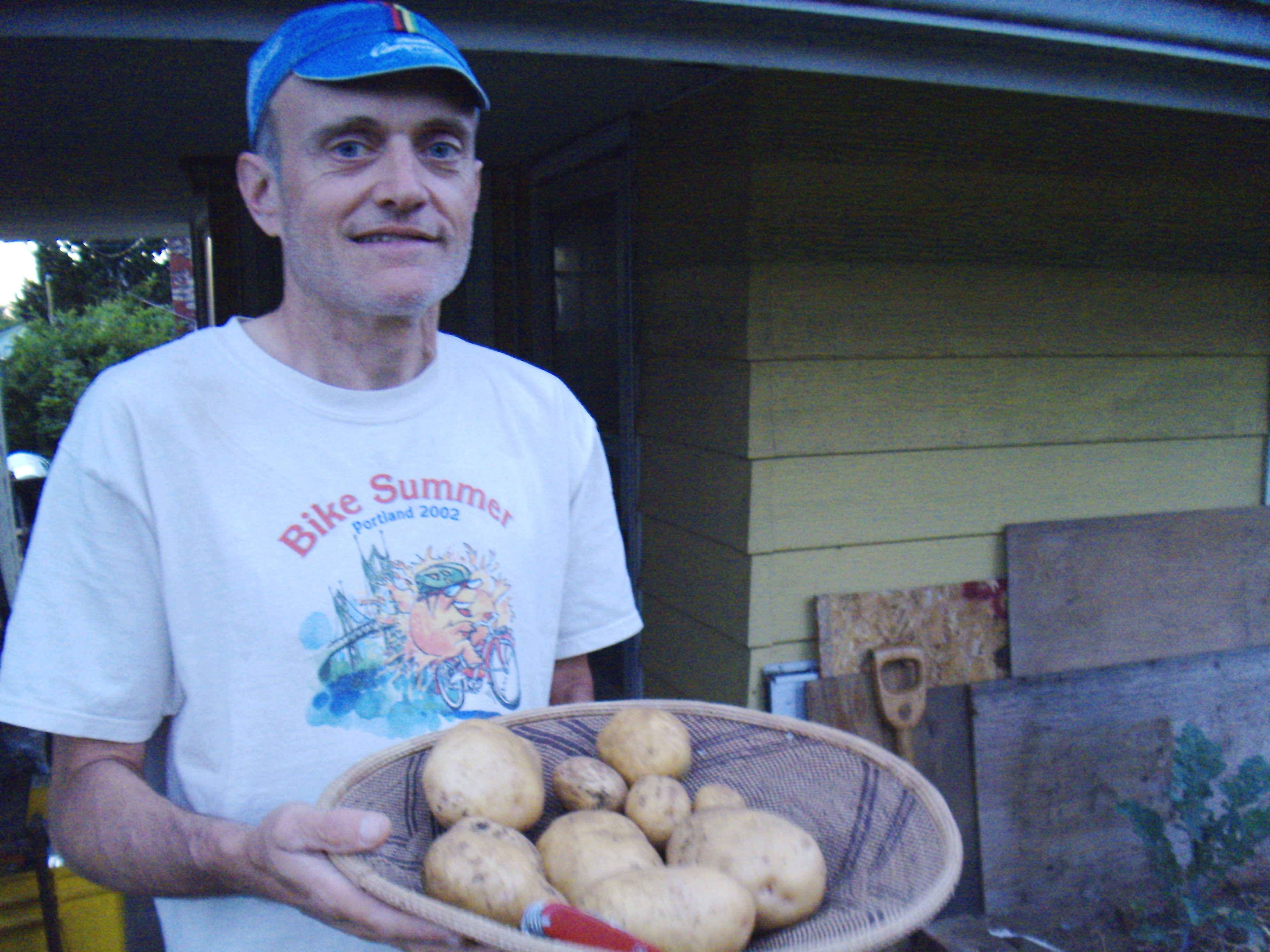 Man holding a basket of potatoes in front of a yellow house.