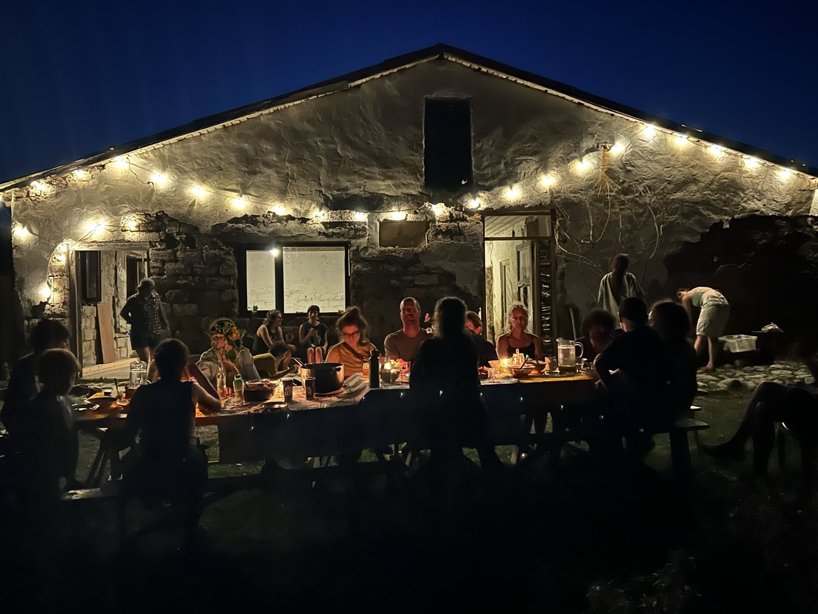People gathering around a long table outside a lit house at night in Ecovillage Georgia.