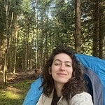 Young woman smiling in front of a blue tent in a forest setting.