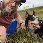 Smiling man with glasses and beard with his dog in a grassy field.
