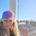 Woman pointing at bridge structure with rainbow-colored cap in bright sunlight.