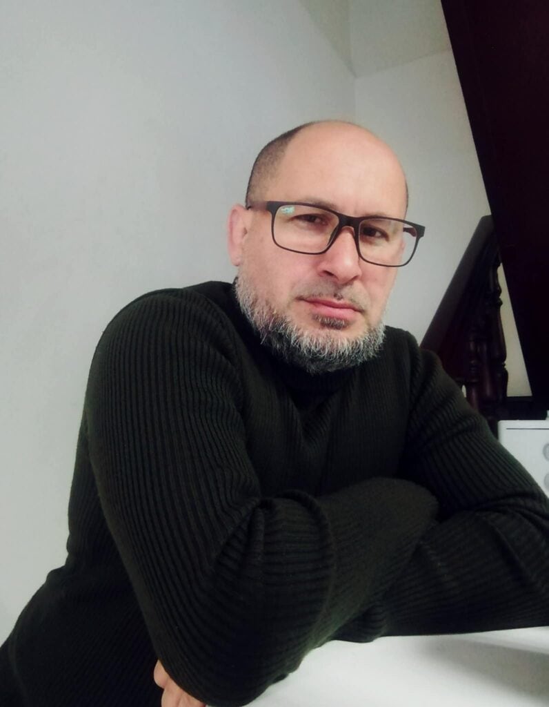 Man with glasses and beard in black sweater at desk.