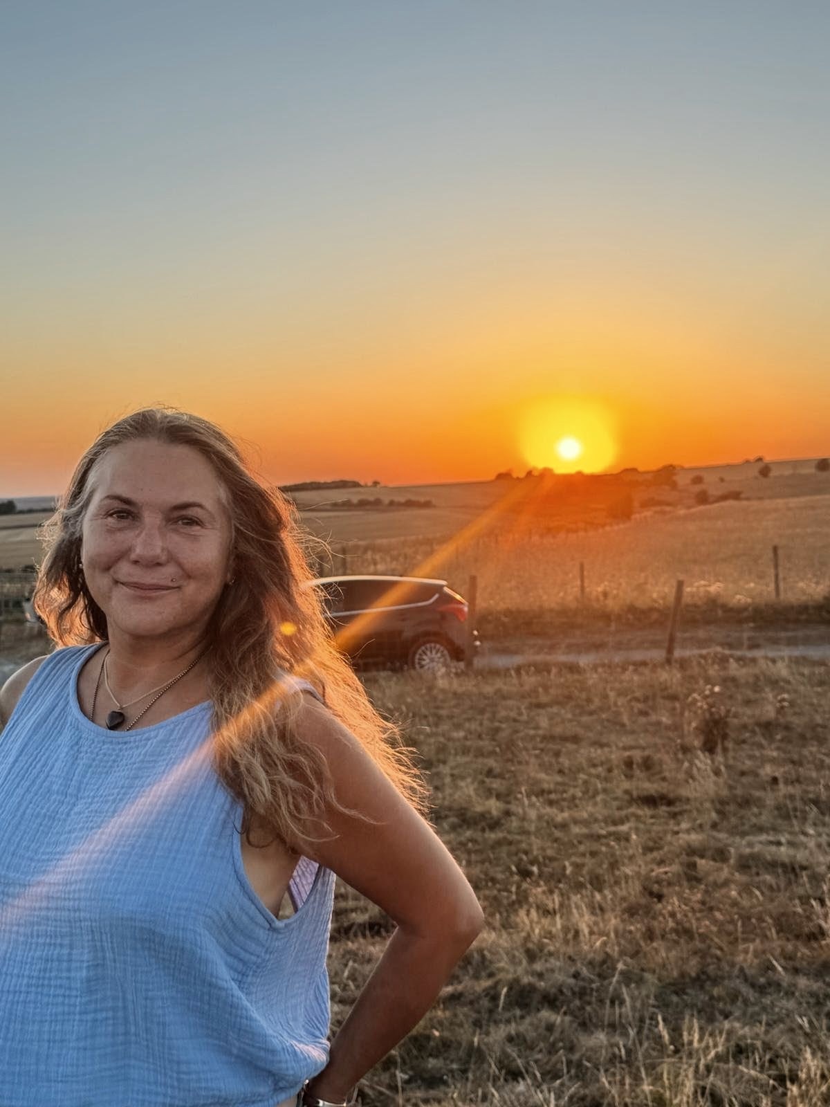 Woman smiling at sunset in a rural community setting with open fields and a car in the background.