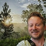 Smiling man in nature with sunrise and trees in background.