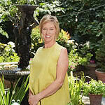Woman smiling in a lush garden with plants and a fountain.