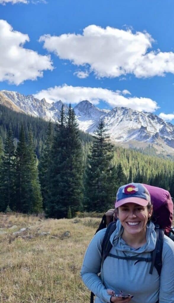 Hiker smiling in mountain landscape with trees and snow-capped peaks.