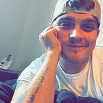 Young man with a cap smiling, resting his face on his hand.