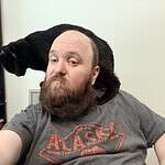 Man with beard and gray T-shirt with black cat on shoulder in community room.