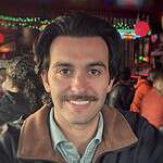 Smiling man at community event with colorful lights and people in background.