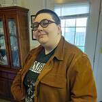 Person with glasses wearing a brown jacket indoors near a window.