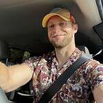 Person in car wearing colorful shirt and cap, smiling, with logo in background.