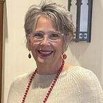 Smiling woman with short gray hair, glasses, red jewelry, and beige sweater.