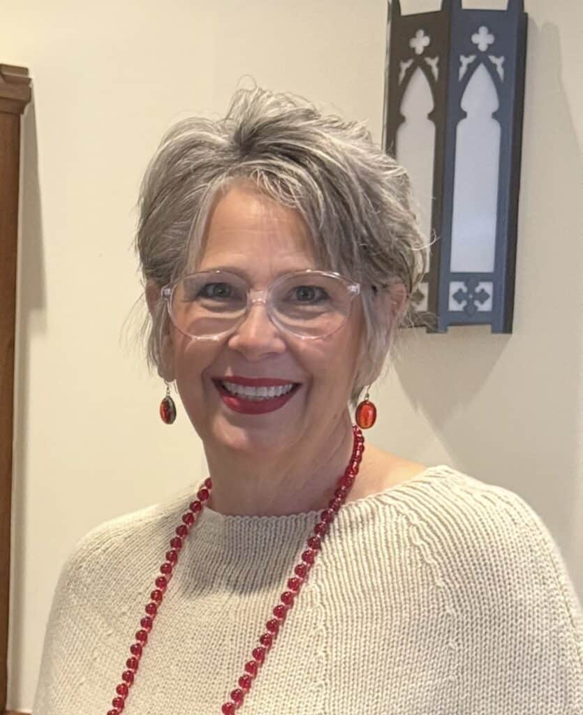 Smiling woman with short gray hair, glasses, red jewelry, and beige sweater.