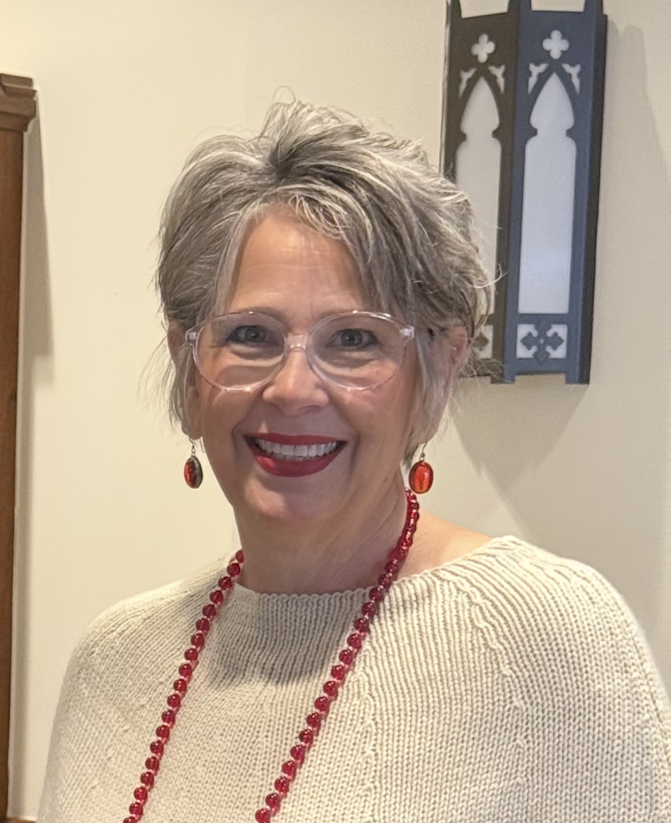Smiling woman with short gray hair, glasses, red jewelry, and beige sweater.