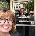 Woman smiling outside Mulligans bar with business hours and ATM sign.
