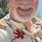 Smiling man with sunglasses outdoors near a lake and mountains.