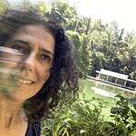 Woman smiling near a lake with lush greenery and modern pavilion in background.