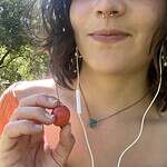 Close-up of a young woman holding a cherry outdoors with sunlight and trees.