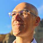 Close-up of a smiling man with glasses outdoors under blue sky.