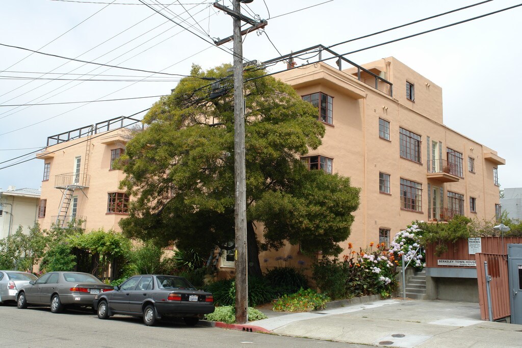 Apartment building with lush tree and parked cars in front.