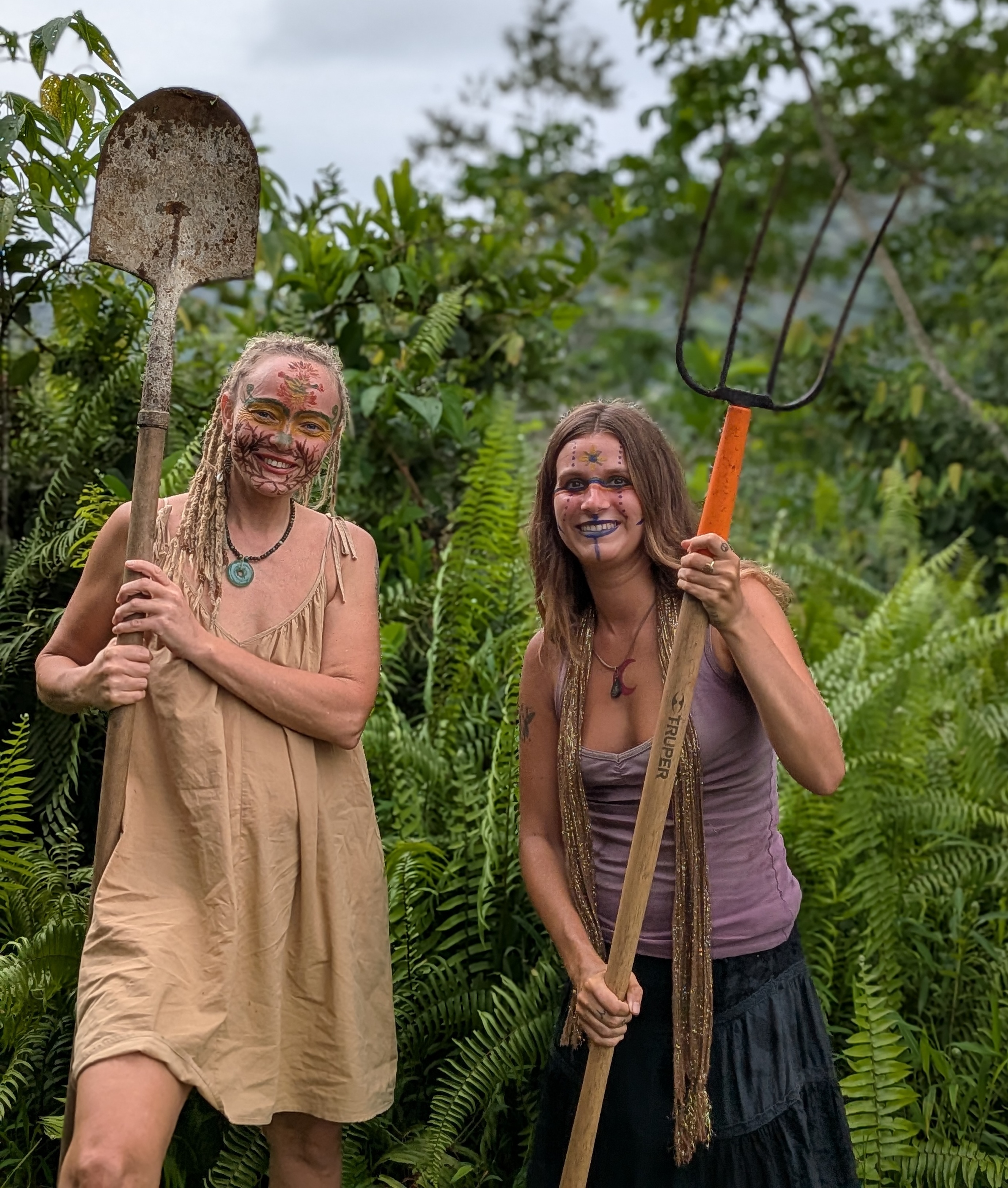 Two women with face paint holding gardening tools in lush greenery.