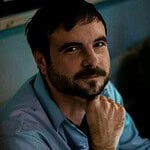 Man with beard and blue shirt, thoughtful expression, in community setting.