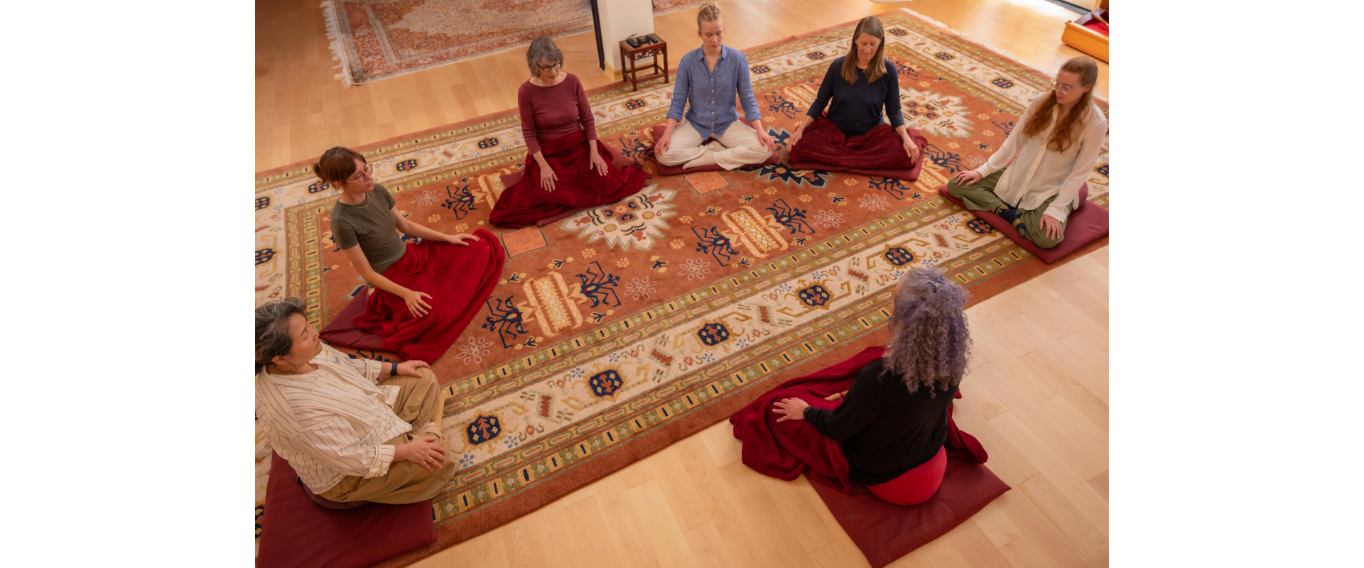 People meditating in a circle on a large rug in a community space.