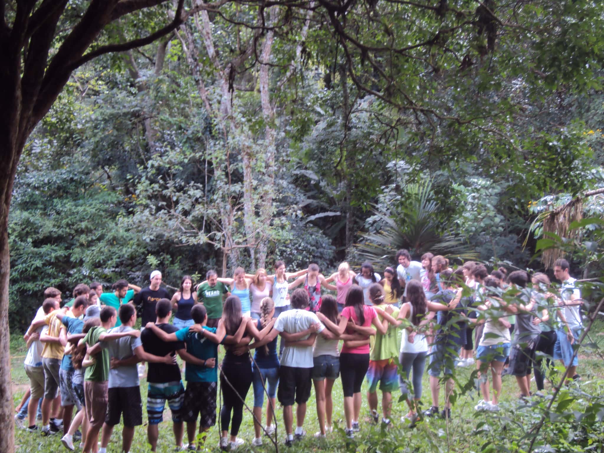 Group of people holding hands in circle in a lush outdoor setting.