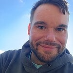 Person smiling outdoors with blue sky and clouds in background.