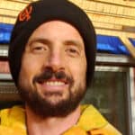 Smiling man wearing a black beanie and yellow jacket outdoors.