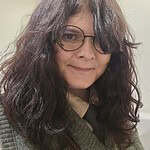 Woman with glasses and wavy hair, smiling, in an indoor setting.