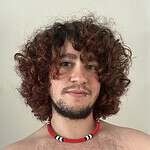Portrait of a young man with curly hair and a beaded necklace, shirtless, against a plain background.