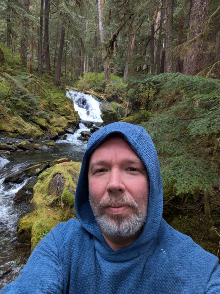 Man in blue hoodie taking selfie near forest stream and waterfall.