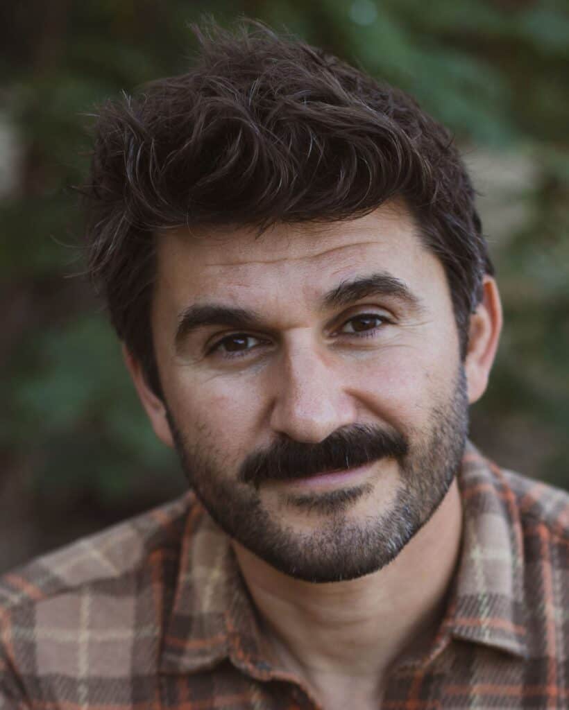 Close-up portrait of a man with dark hair and beard outdoors.